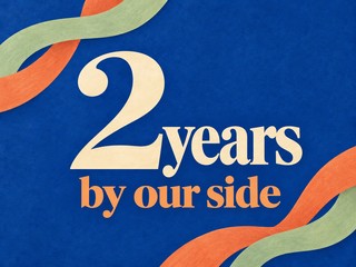 Illustration of cream and apricot lettering with colorful ribbons on cobalt blue, with the message “2 years by our side”